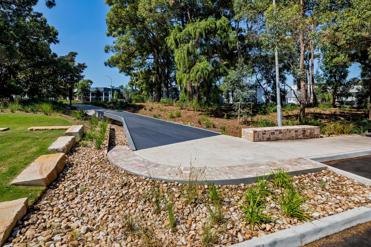 Paved park entrance with stone seating blocks, river pebble garden beds and eucalyptus trees in Sydney Park.