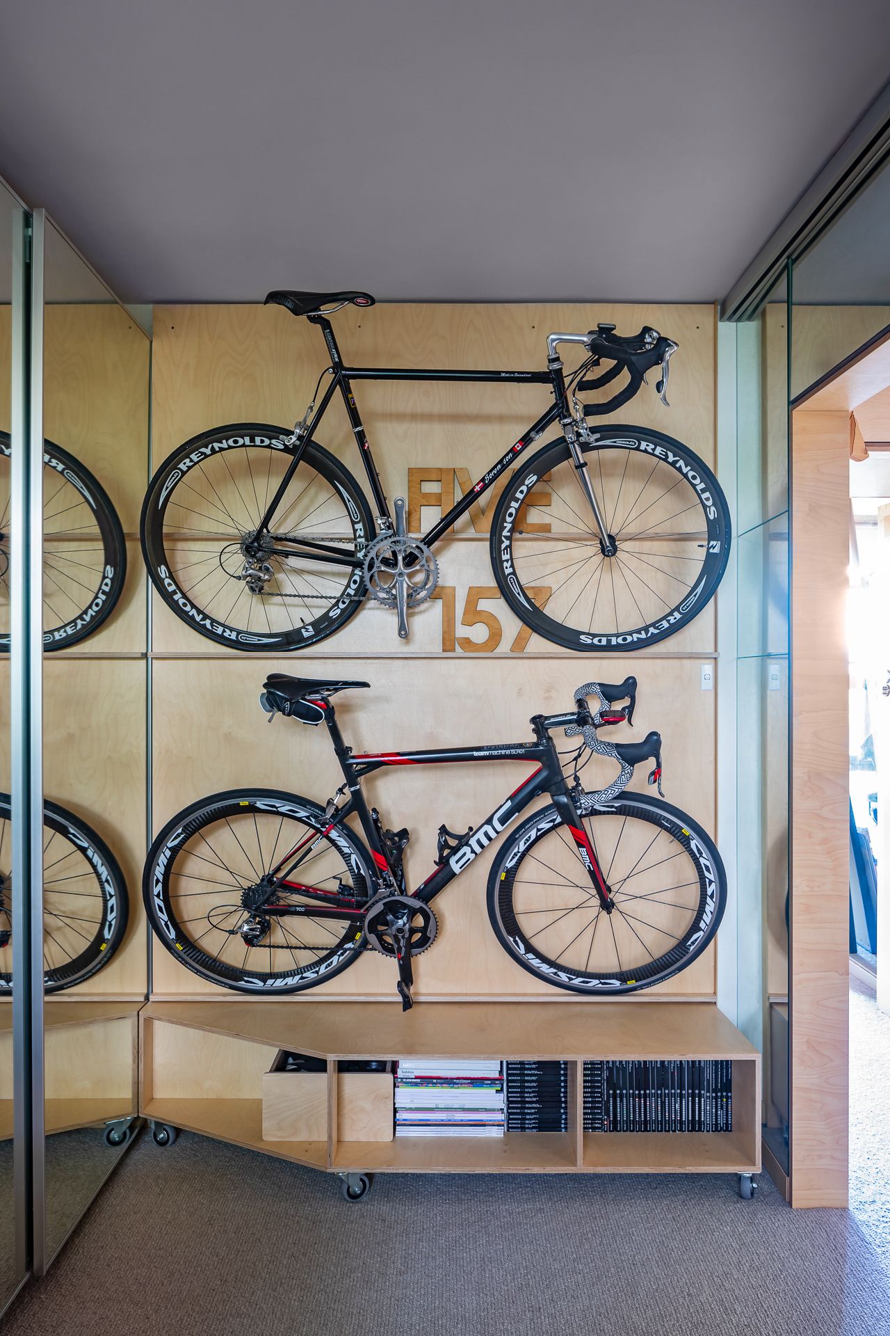 Two road bikes mounted on a plywood wall above a rolling storage shelf with books, beside a mirrored sliding door.