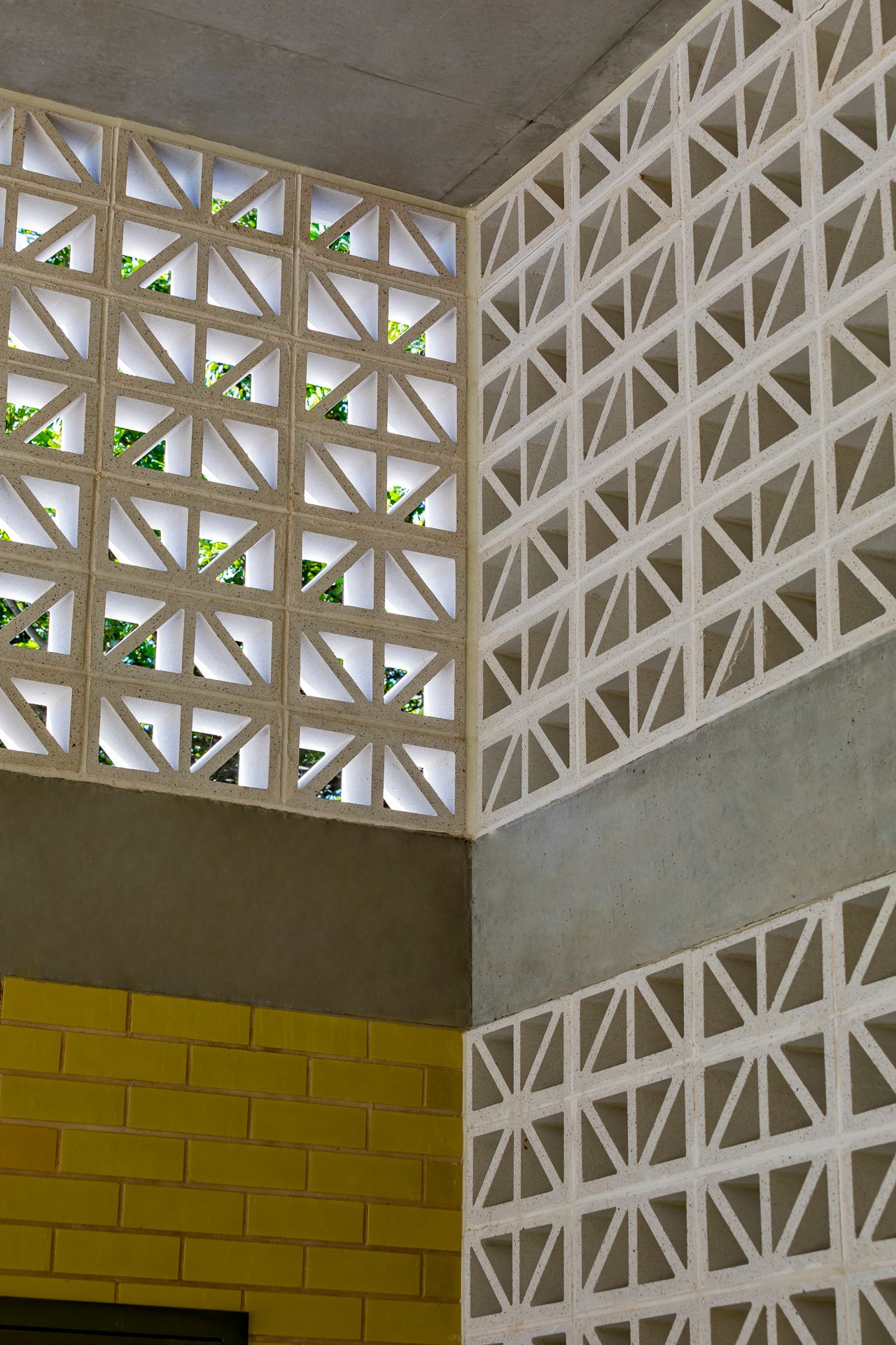 Close-up of white geometric breeze block walls meeting yellow brick and concrete, with green foliage visible through the openings.