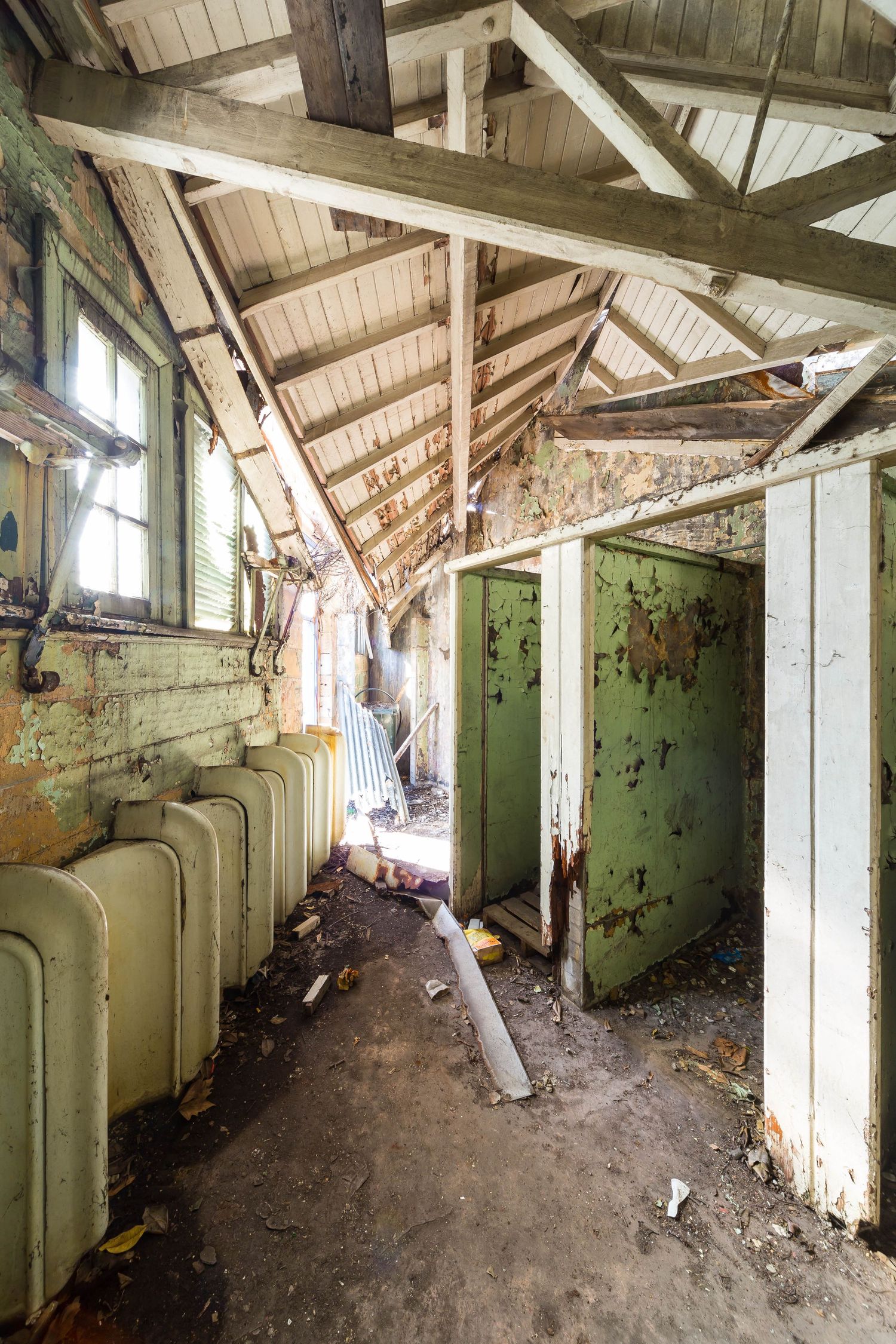 Derelict building interior with exposed timber roof trusses, peeling green paint, old fixtures and debris on the ground.