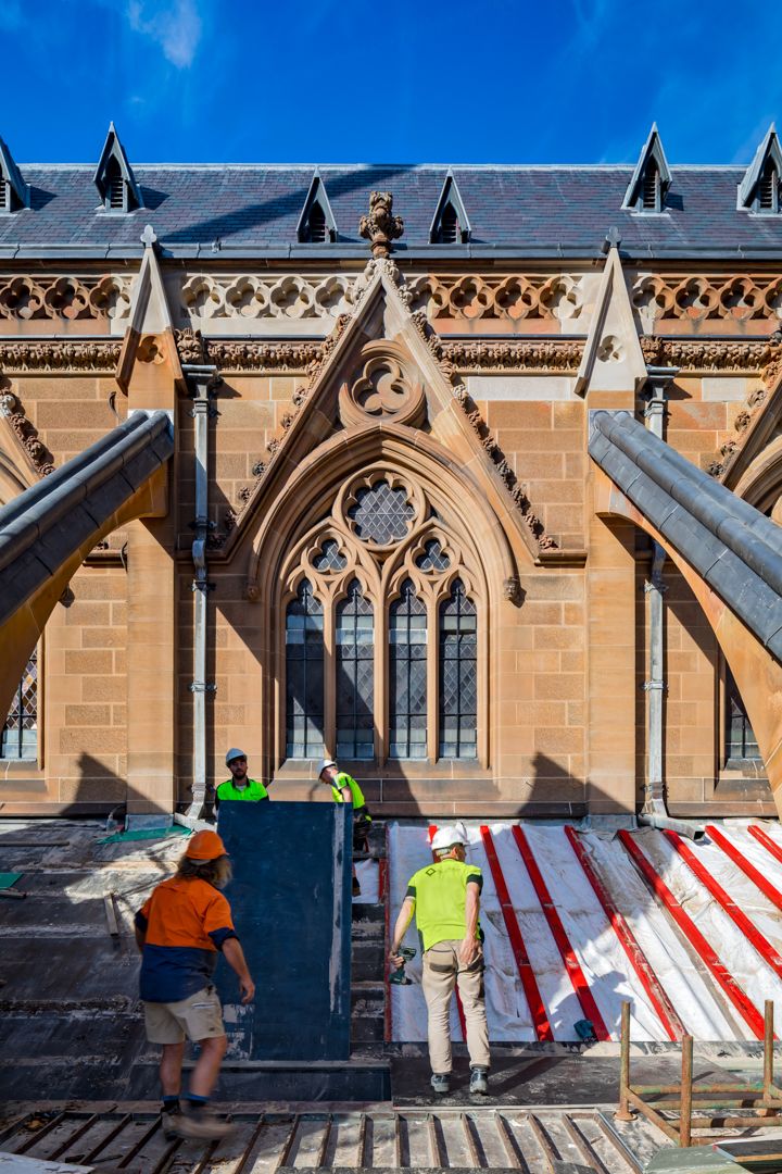 Workers in hi-vis clothing carry out roof restoration beside an ornate sandstone Gothic building with arched windows.