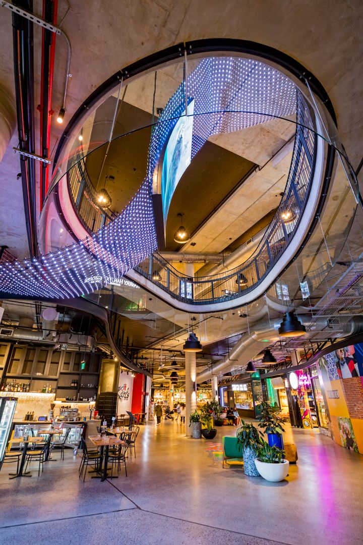 Modern shopping and dining atrium with a circular skylight opening, LED light installation, cafe tables and potted plants.