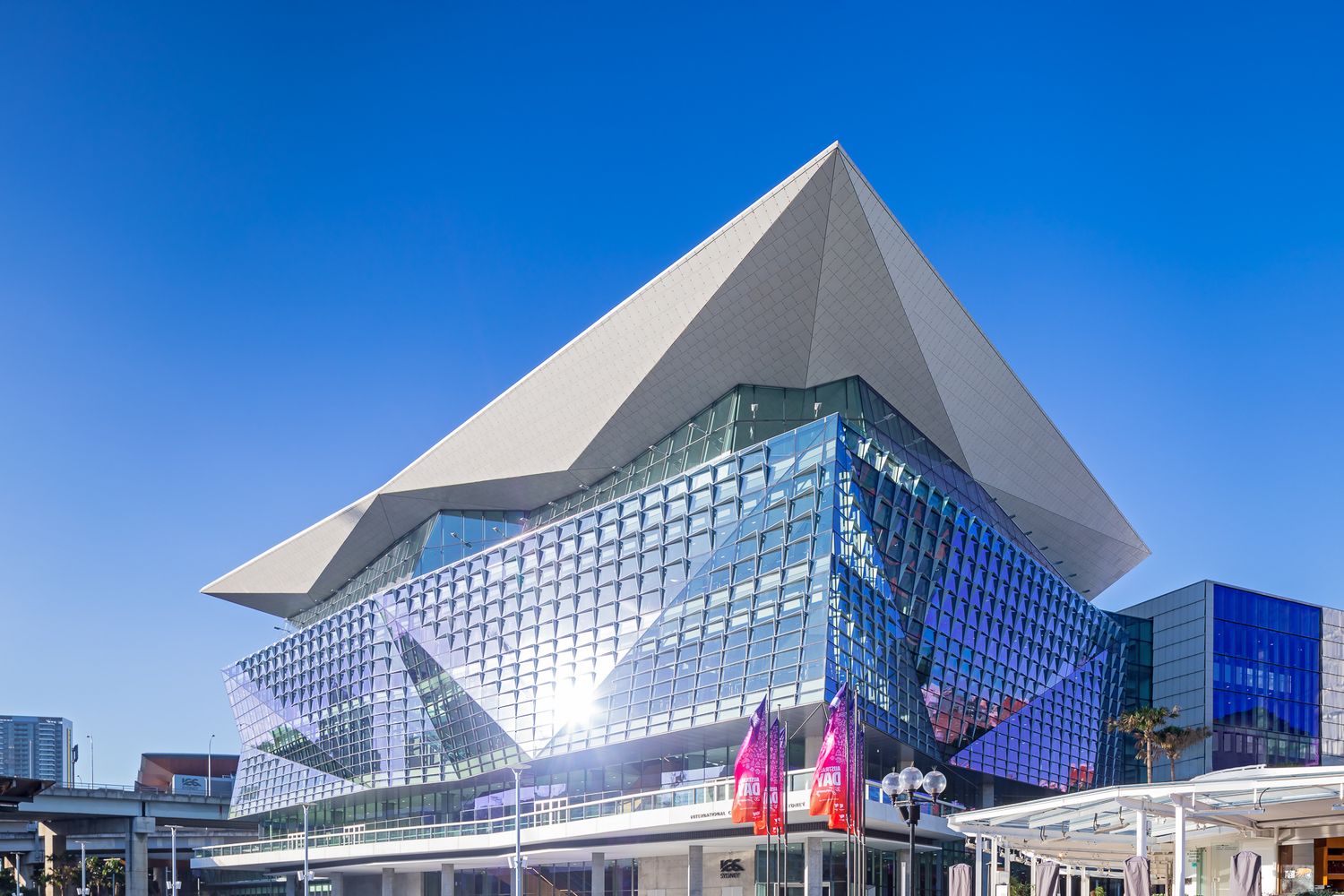 Contemporary glass convention centre with a dramatic pyramid-shaped roof, colorful banners and a clear blue sky.