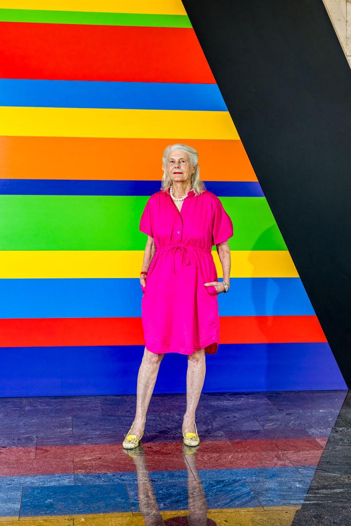 An older woman in a bright pink dress stands in front of a colorful striped wall mural, with her reflection visible on the glossy floor.