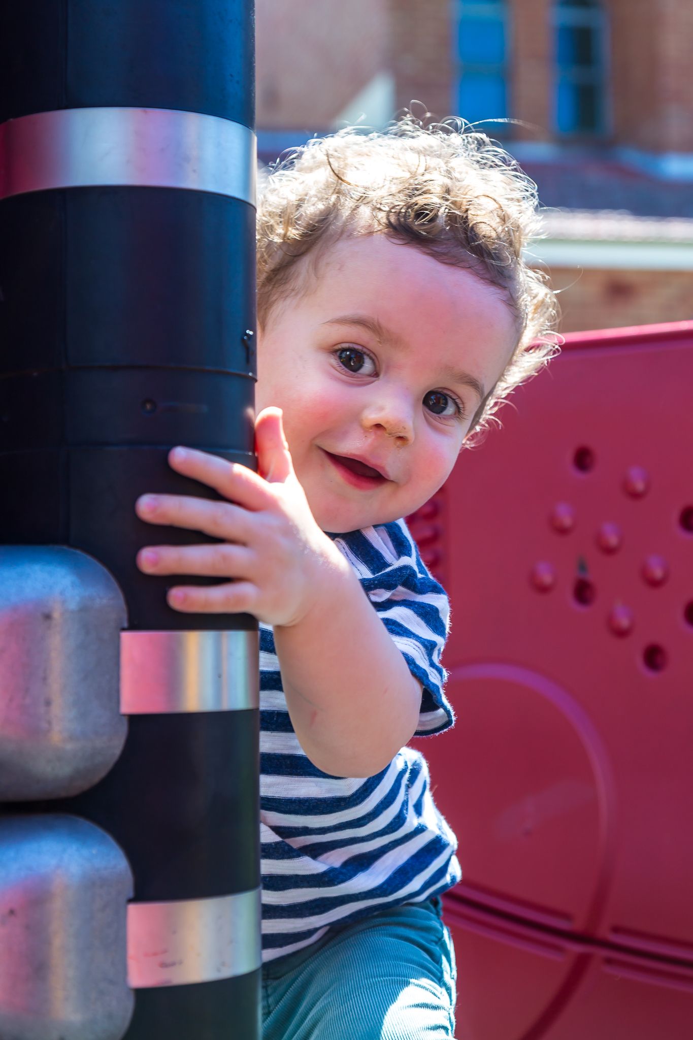 Smiling curly-haired toddler in a striped shirt peeks around a black pole on a sunny playground.