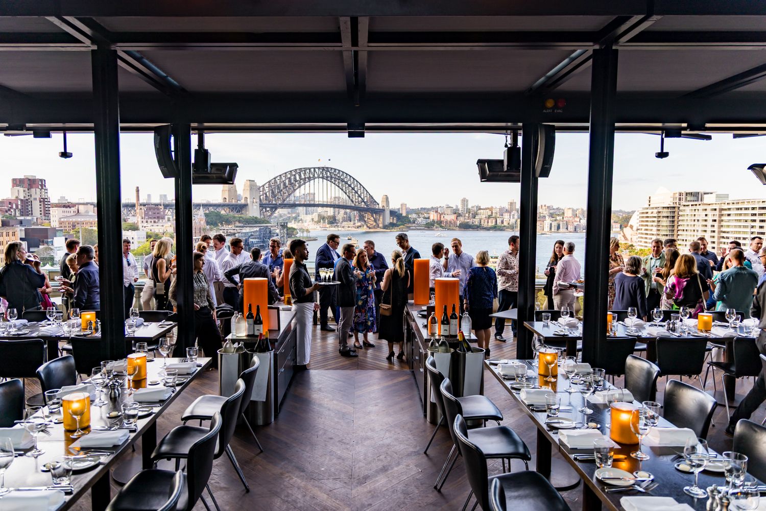 Guests gather at a waterfront restaurant terrace with a view of the Sydney Harbour Bridge at golden hour.