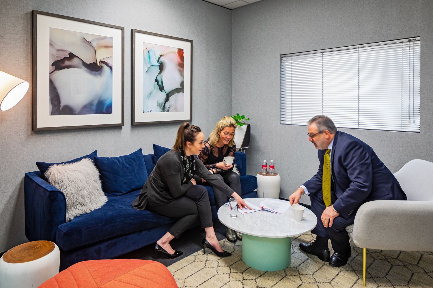 Two women and a man in a suit talk around a marble coffee table in a modern office lounge with a blue sofa and framed art.