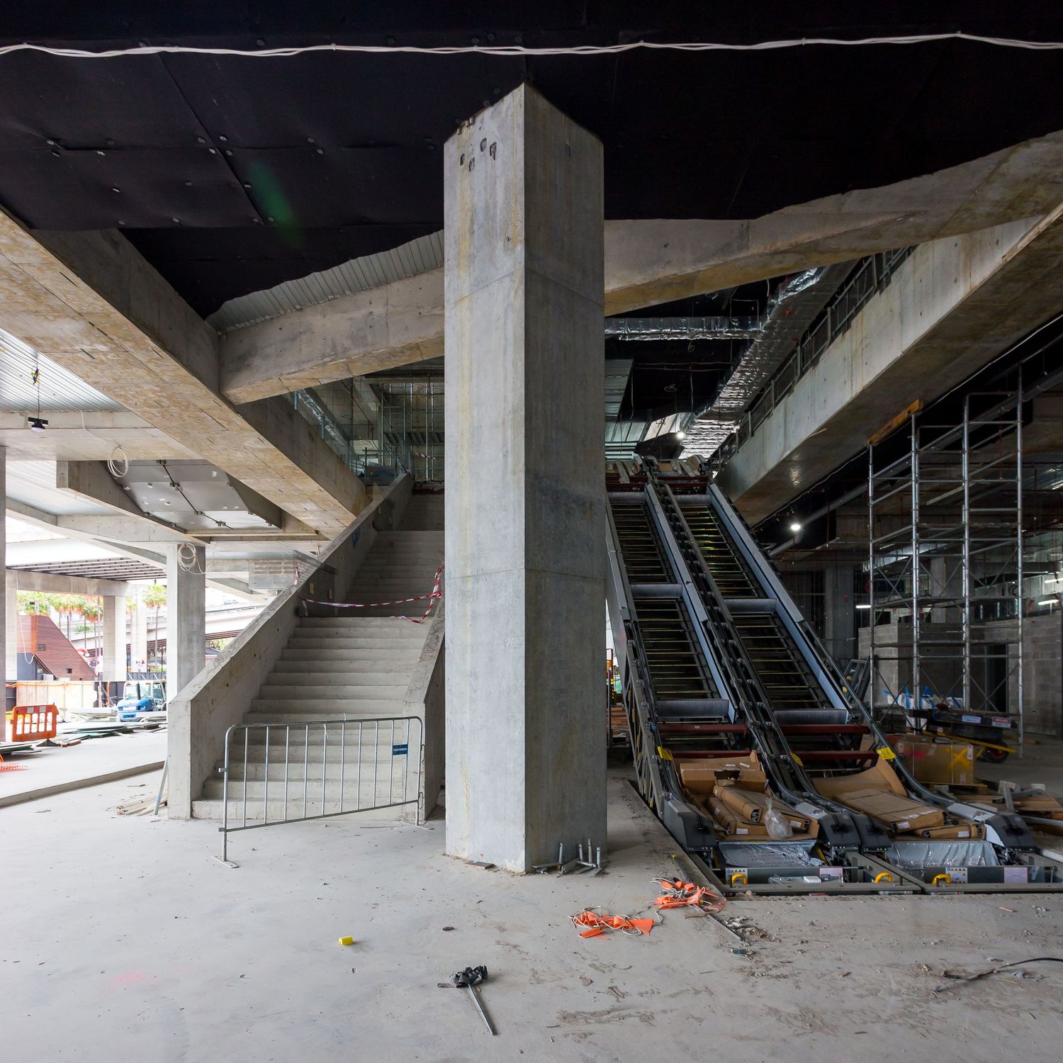 Concrete columns and beams surround partially installed escalators in a large building under construction with protective barriers.