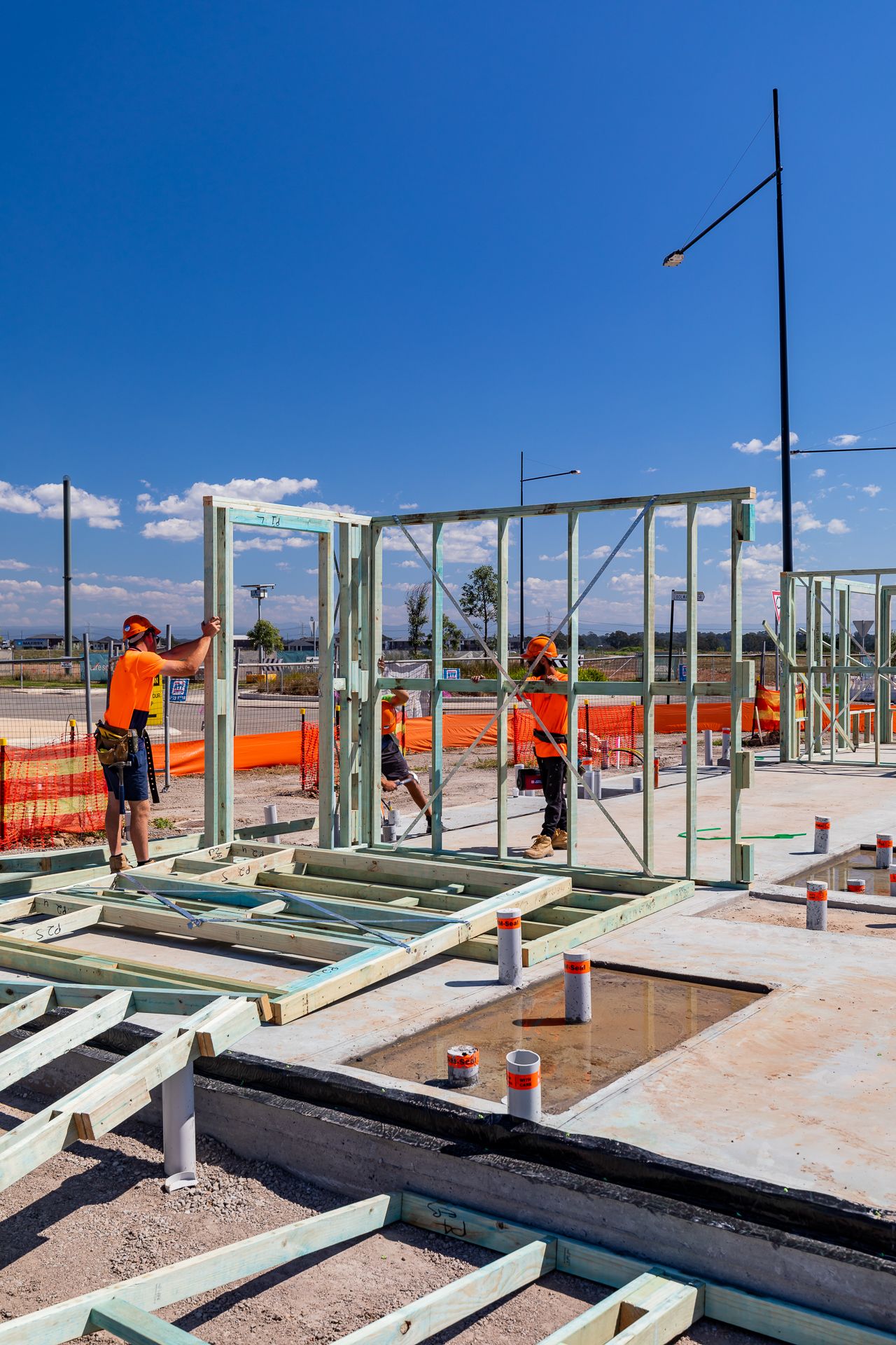 Tradespeople in orange hi-vis clothing erecting steel wall frames on a concrete slab at a residential construction site under blue sky.