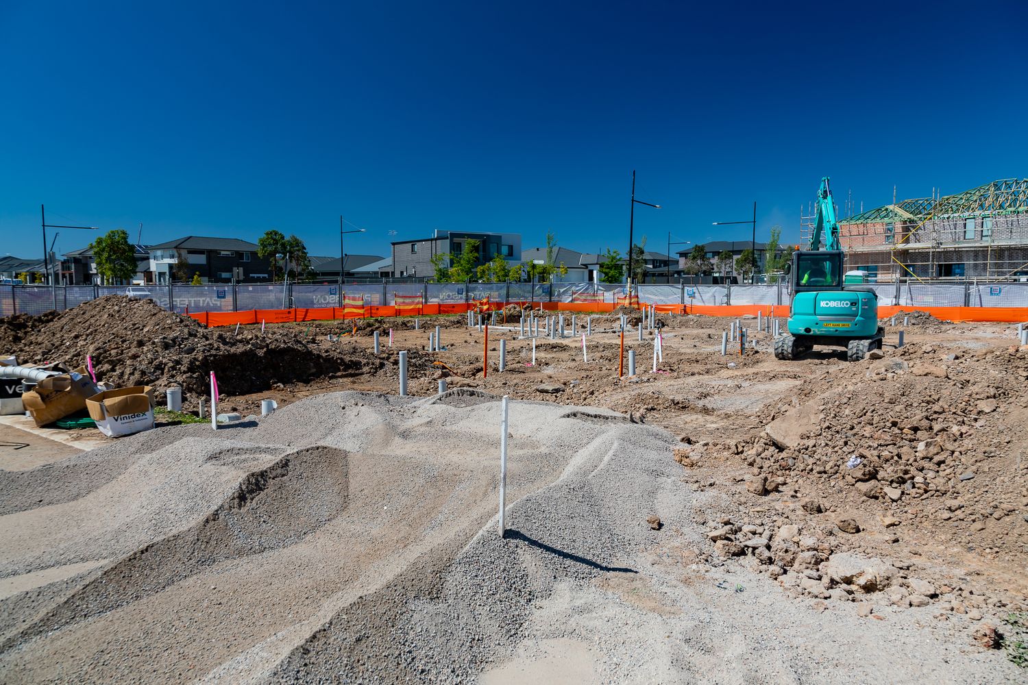 Excavator and mounds of soil on a residential construction site with orange safety fencing and houses in the distance.
