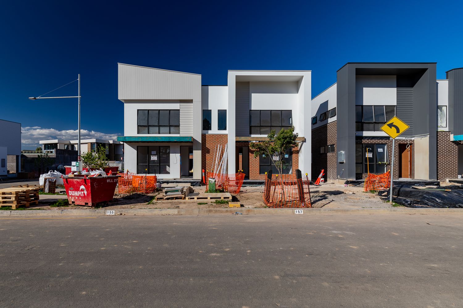 Modern townhouses with brick and render facades under construction on a street with orange safety fencing and a waste skip bin.