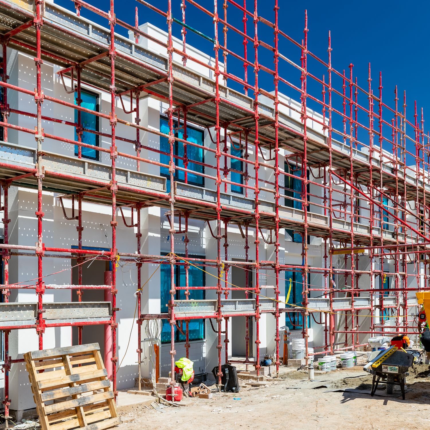 Multi-storey residential building under construction wrapped in red scaffolding with workers and timber pallets in the foreground.