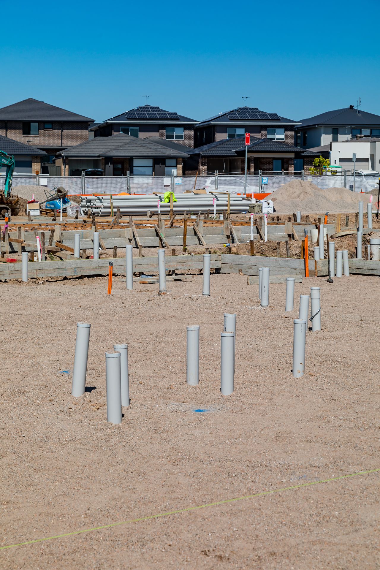 Grey plumbing pipes stand upright in bare earth at a residential construction site with finished homes in the background.