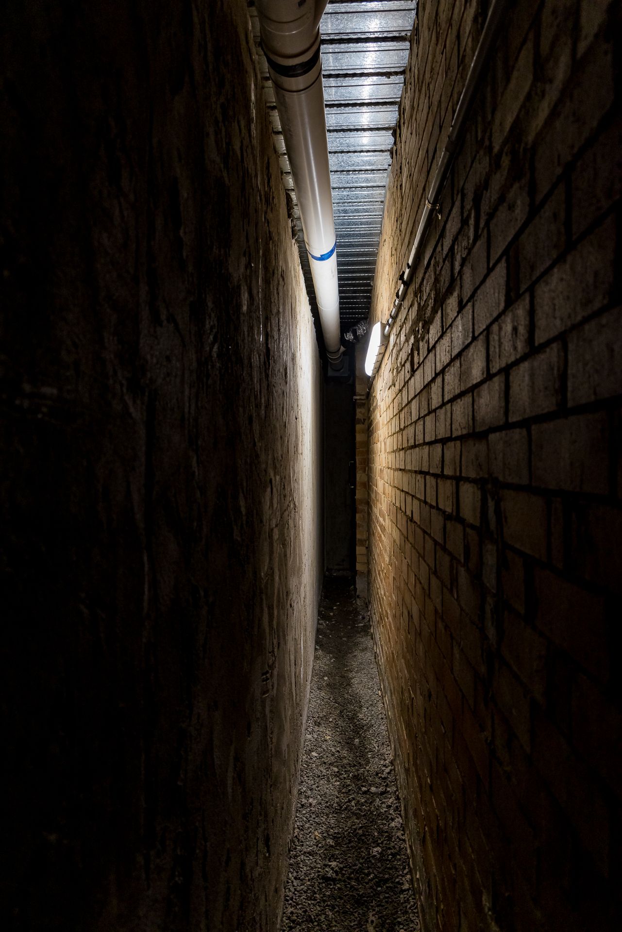 Narrow gravel passage between two tall brick walls lit by a single light, with a pipe running along the ceiling.