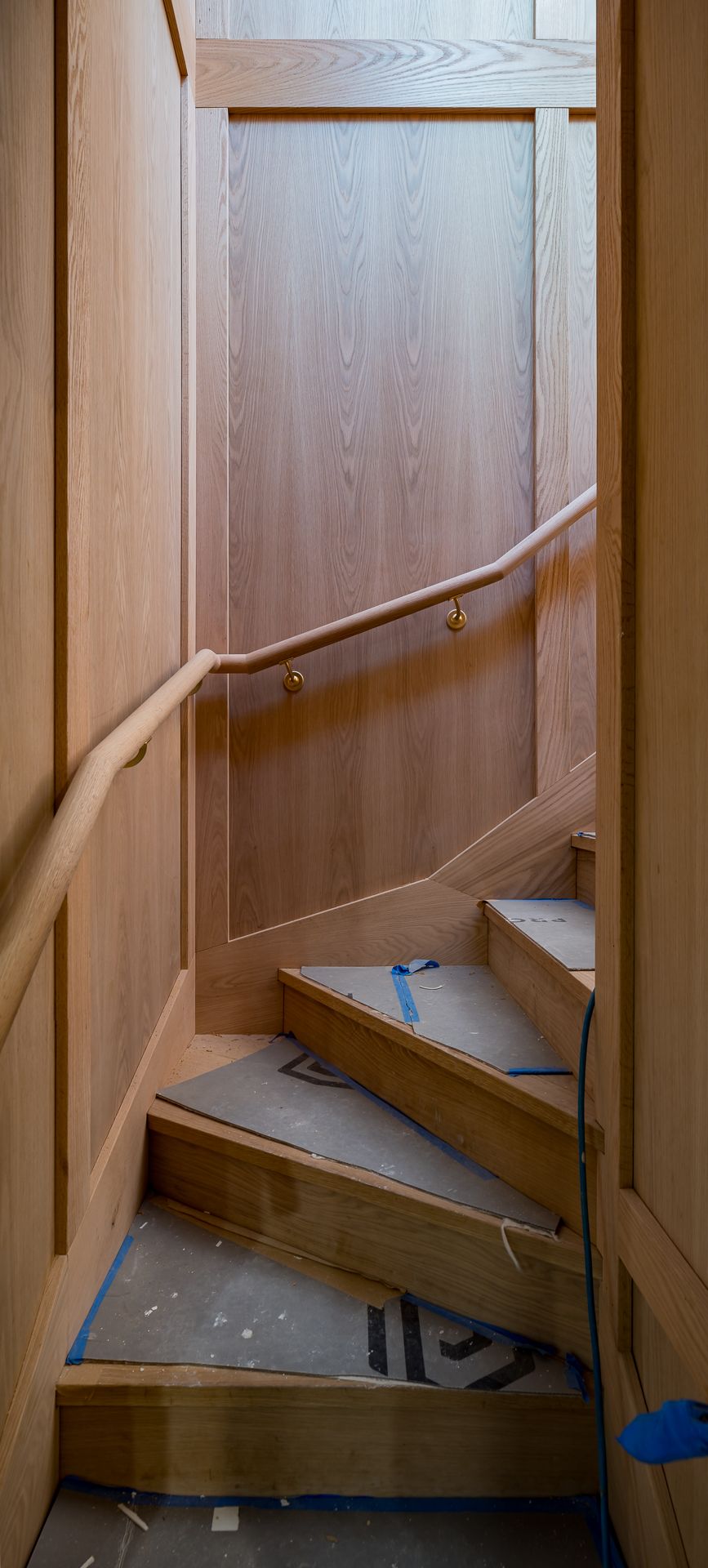Narrow timber-paneled staircase with a curved wooden handrail and protective covering on the steps during an interior home renovation.
