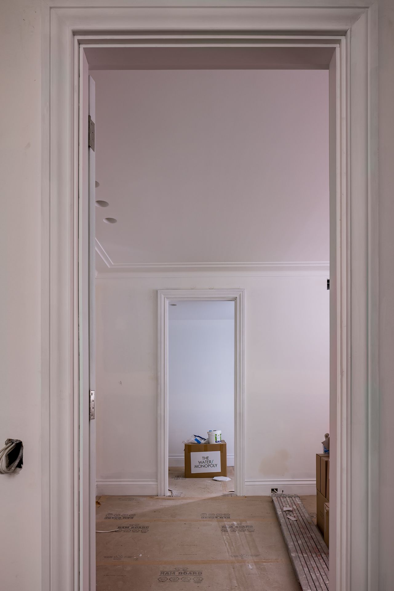 View through two white-trimmed doorways into an unfinished renovated room with a cardboard box and paint tin on the floor.