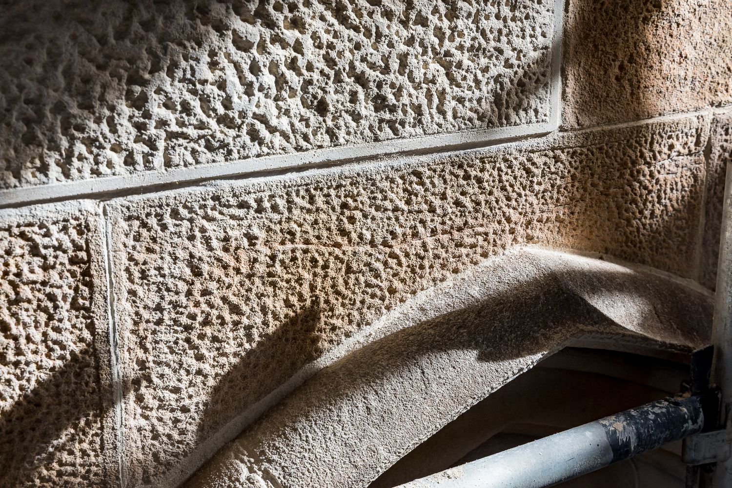 Weathered limestone wall with rough pitted texture, smooth carved edges and a metal pipe at the lower right.