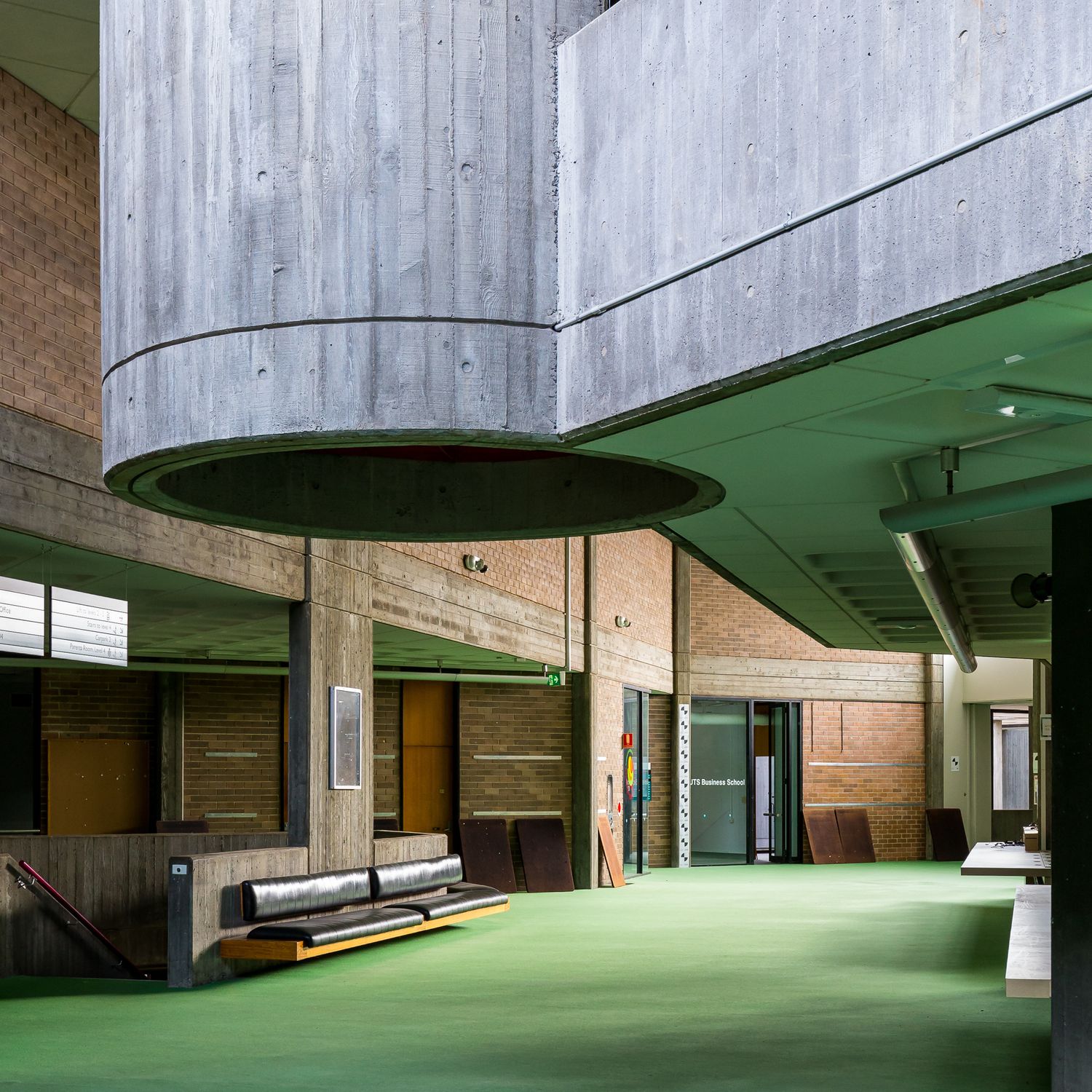 Brutalist interior with a large cylindrical concrete form, exposed brick walls, timber bench seating and green carpeted floor.