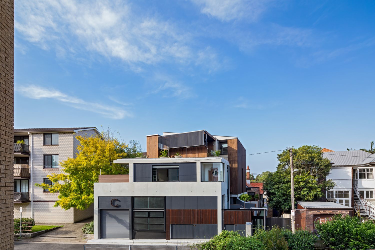 Contemporary multi-level townhouse with dark cladding and a garage, set beside a brick apartment block and a leafy autumn tree.