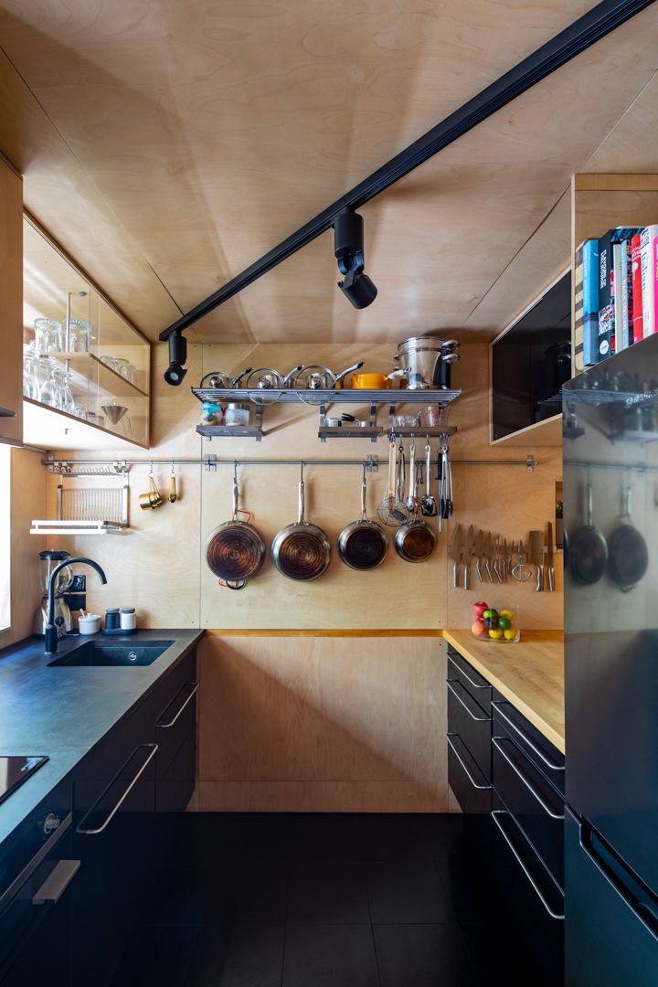 Compact galley kitchen with plywood walls, black cabinetry, hanging copper pots and utensils on a rail, and track lighting overhead.