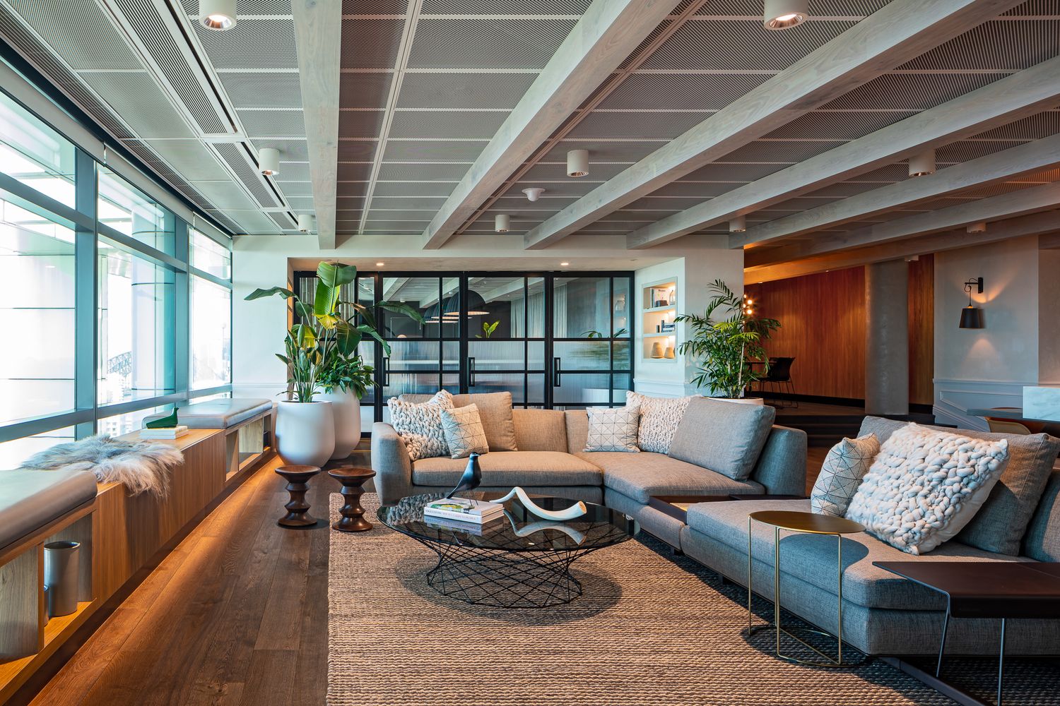 Stylish lounge with gray sectional sofa, patterned cushions, woven rug, indoor plants and exposed beam ceiling beside large glass windows.