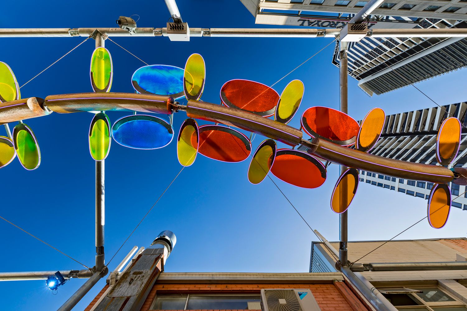 Overhead view of a wind-driven public art sculpture with translucent orange, blue and green discs above a brick building.