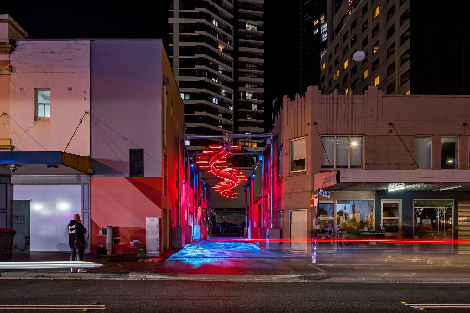 Red neon spiral sculpture glowing above a city laneway entrance at night with high-rise apartment towers in the background.
