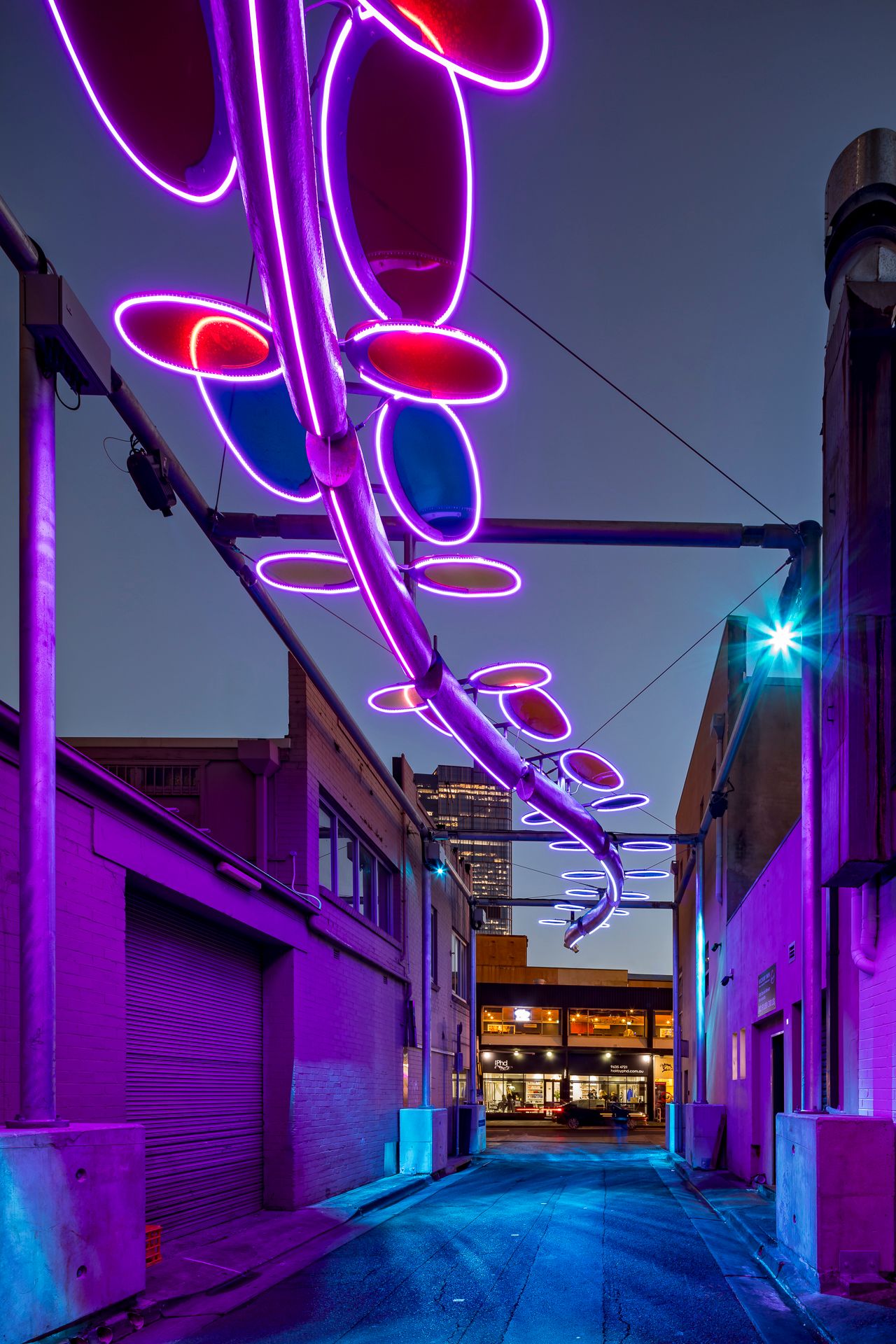 Purple neon-lit serpent sculpture with oval discs suspended over a quiet urban alley at dusk.