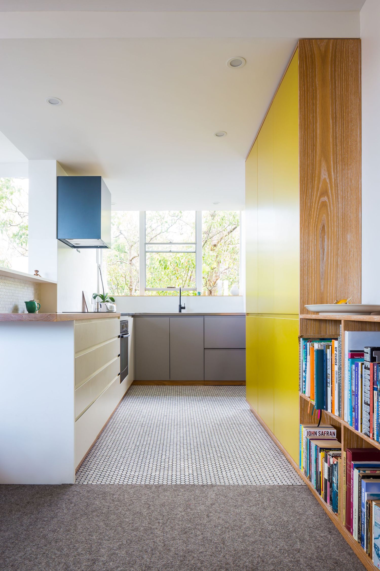 Modern kitchen with yellow cabinetry, timber panels, a blue range hood and a bookshelf leading toward a bright garden window.