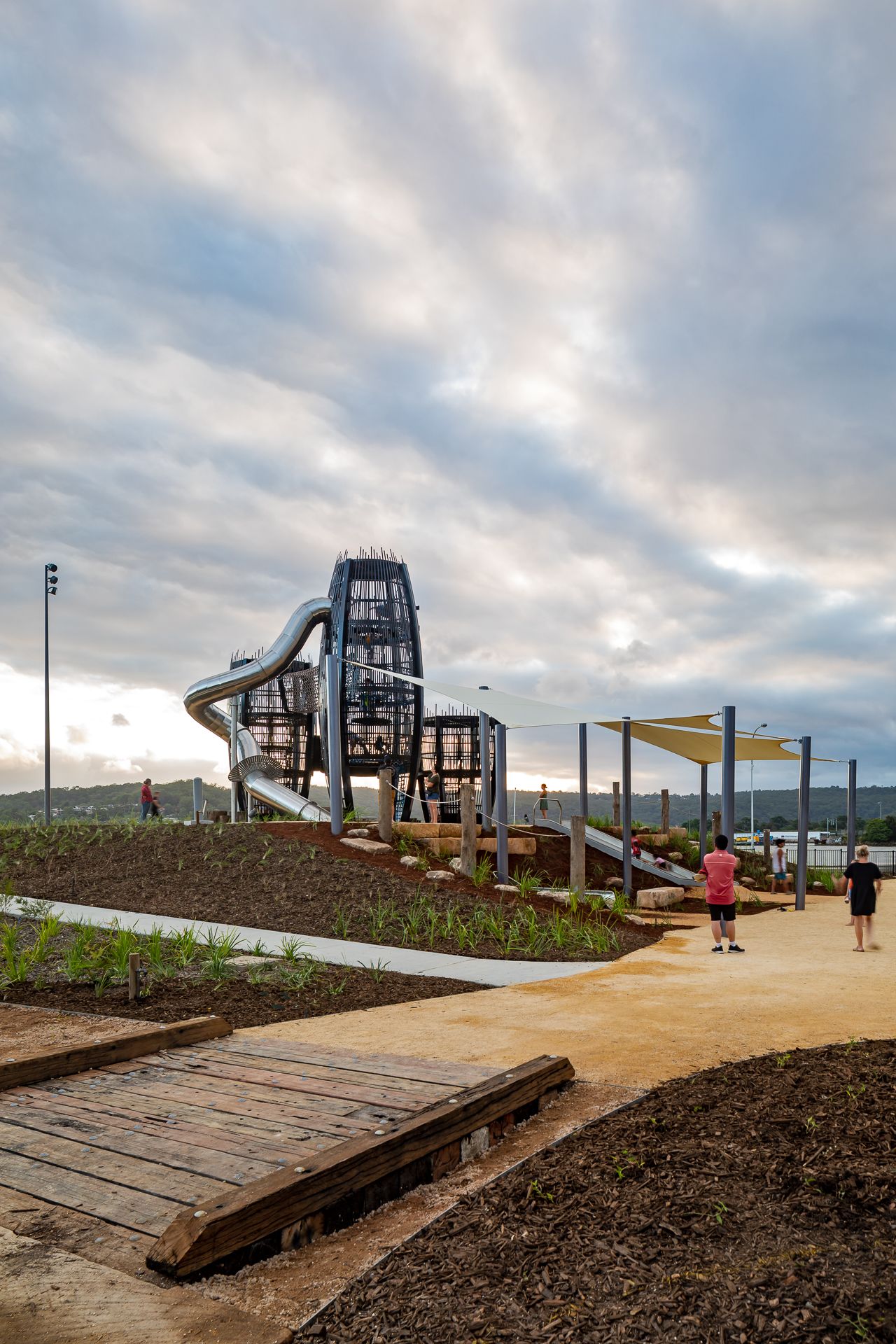 Children's playground with towering net-filled climbing towers and a silver spiral slide beneath a dramatic cloudy sky.