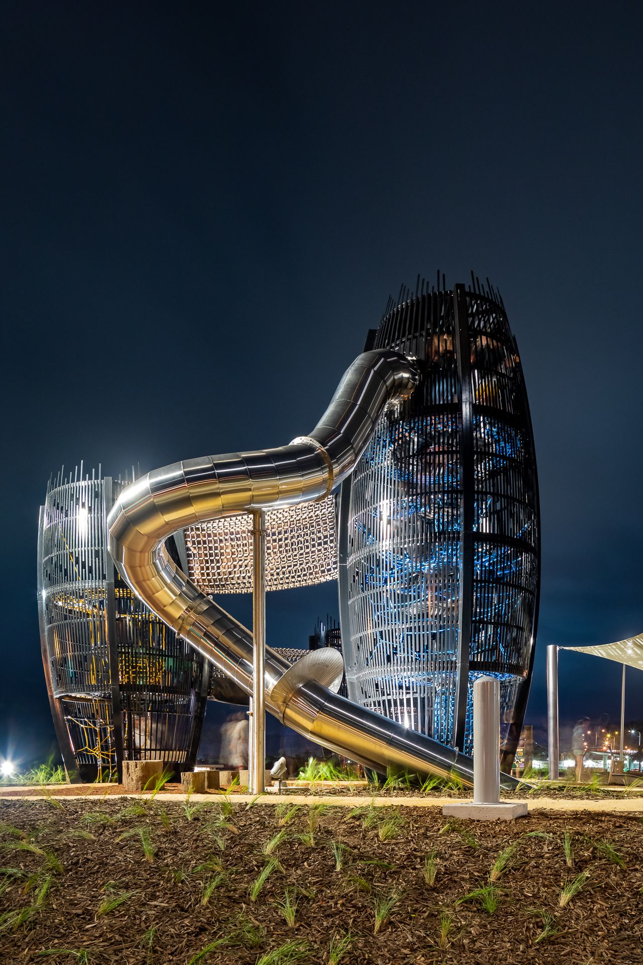 Night view of a giant stainless steel spiral slide winding between two lattice playground towers lit against a dark blue sky.