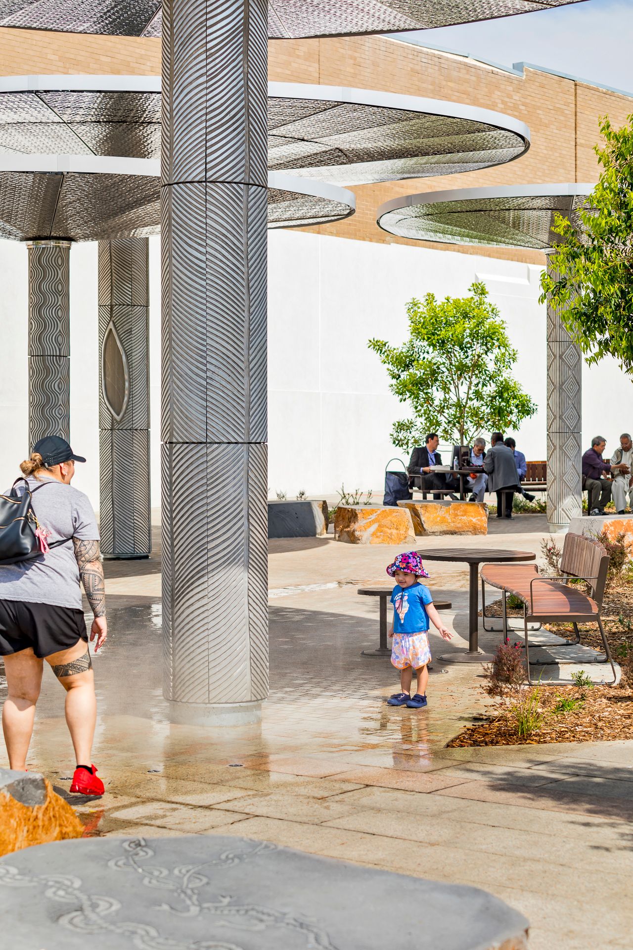 A toddler splashes in a shallow water feature beneath decorative metal shade canopies while adults sit at tables in the plaza.
