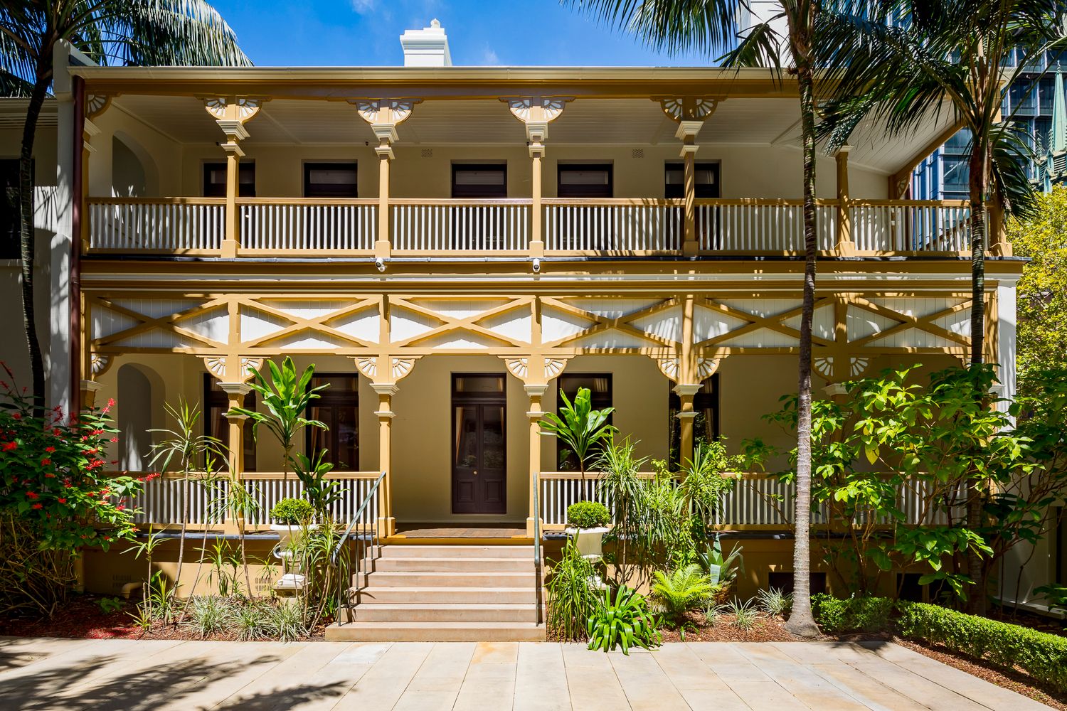 Heritage terrace facade with an upper balcony, ornate timber brackets and a central staircase surrounded by palms and tropical plants.