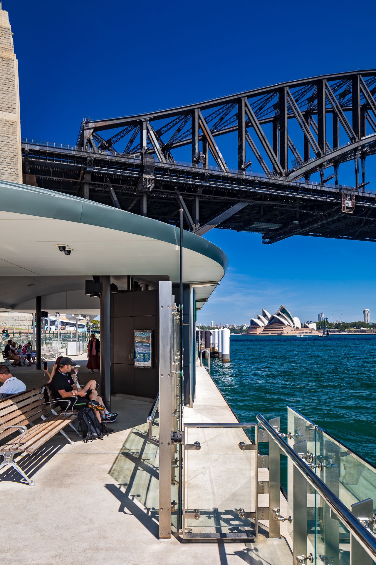 Commuter sits on a bench at a harbourside ferry wharf beneath the Sydney Harbour Bridge with the Opera House in the distance.