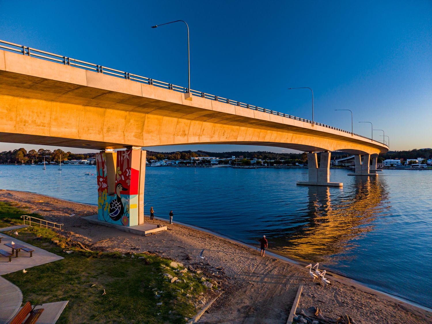 Concrete road bridge with a colorful mural on its pier spanning calm waterfront waters under a clear blue sky at golden hour.