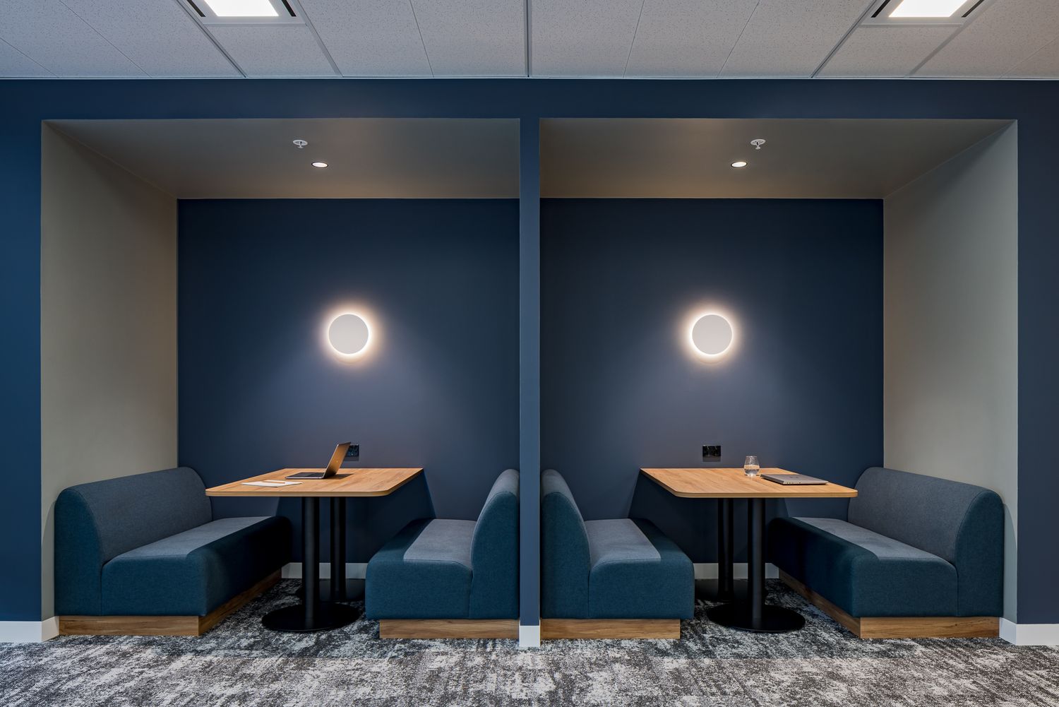 Two private office booths with navy blue banquette seating, wooden tables and round wall lights in a modern workspace.