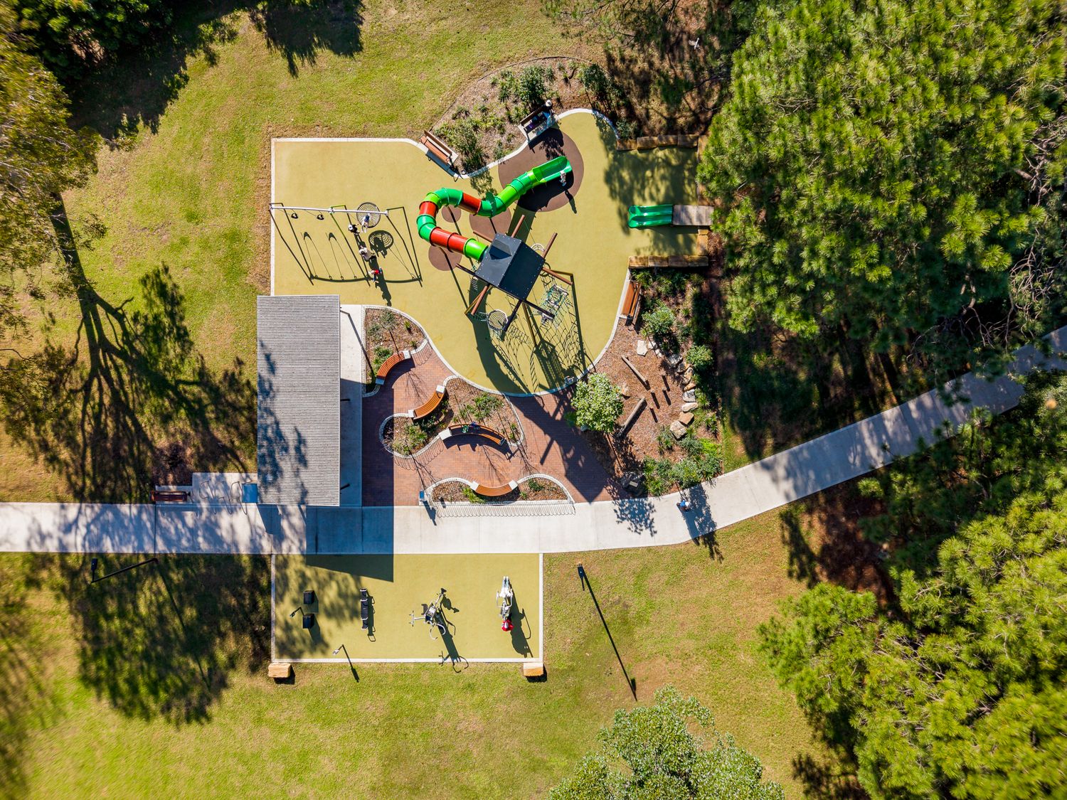 Aerial view of a park playground with green slides, climbing equipment, rubber surfacing and a concrete path surrounded by lawn and trees.
