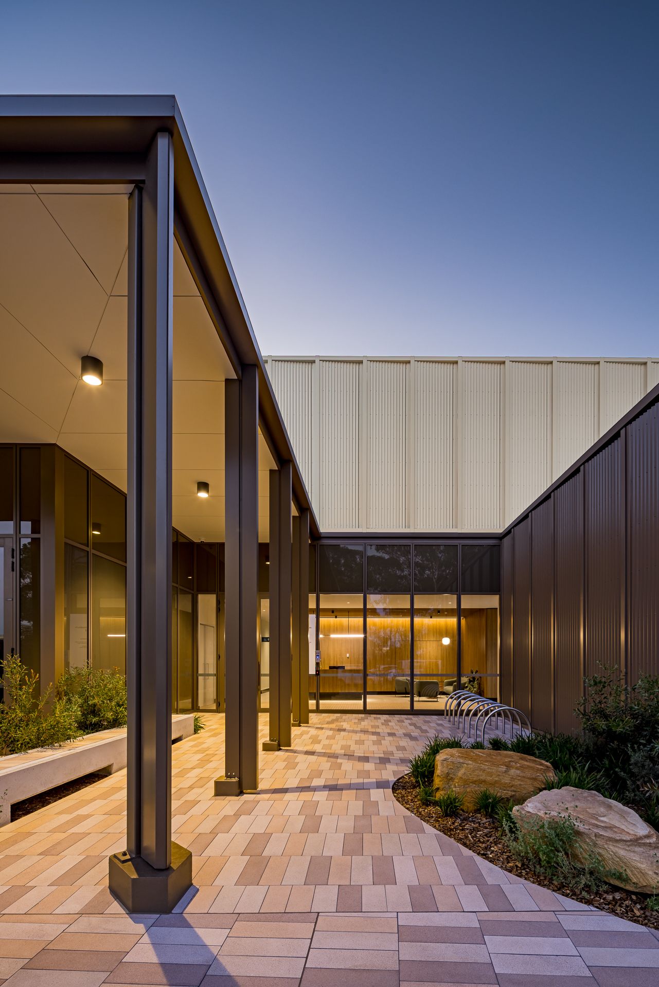 Modern building courtyard at dusk with a covered walkway, steel columns, paved path, native plantings and large boulders near glass doors.