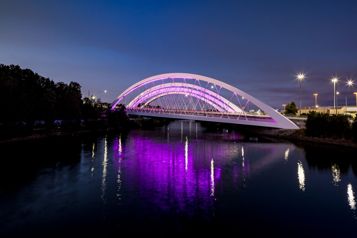 White arch bridge glowing with purple lights at night, reflected in the still river below.