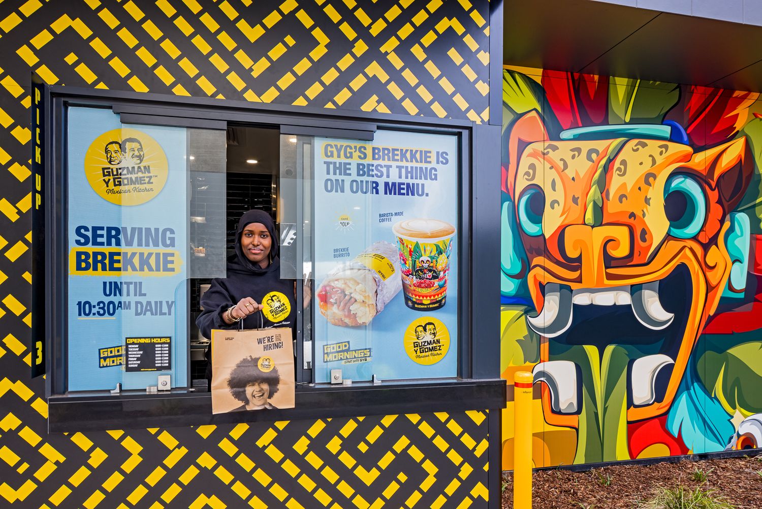 Smiling staff member at a drive-through window serving breakfast beside a colourful Aztec-style mural and yellow pattern wall.