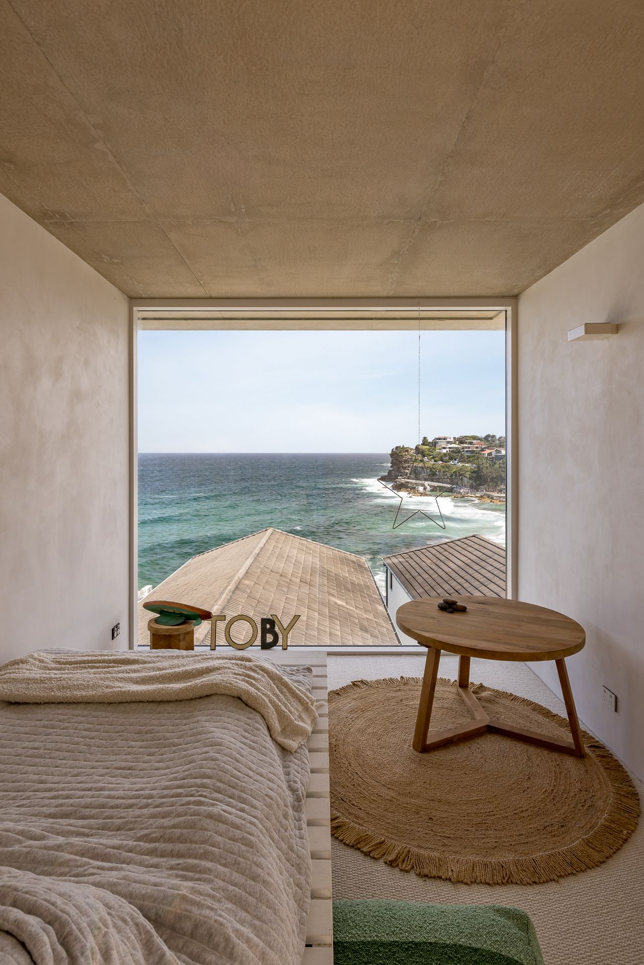 Serene bedroom with a wide ocean-view window, round timber side table, jute rug and textured knit bedding under a concrete ceiling.