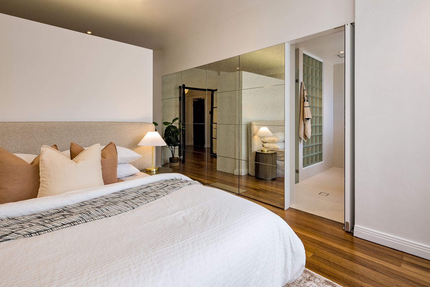Bedroom with a plush bed and neutral cushions, mirrored sliding doors open to reveal an ensuite with glass block walls.