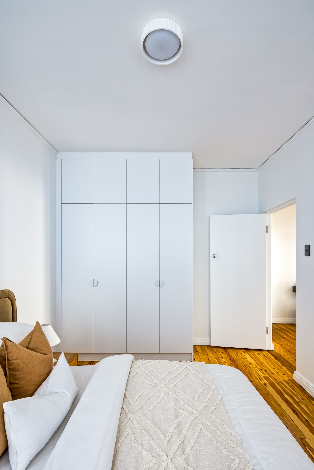Bright modern bedroom with built-in white wardrobes, timber floorboards, a neutral bed with tan cushions and an open door.