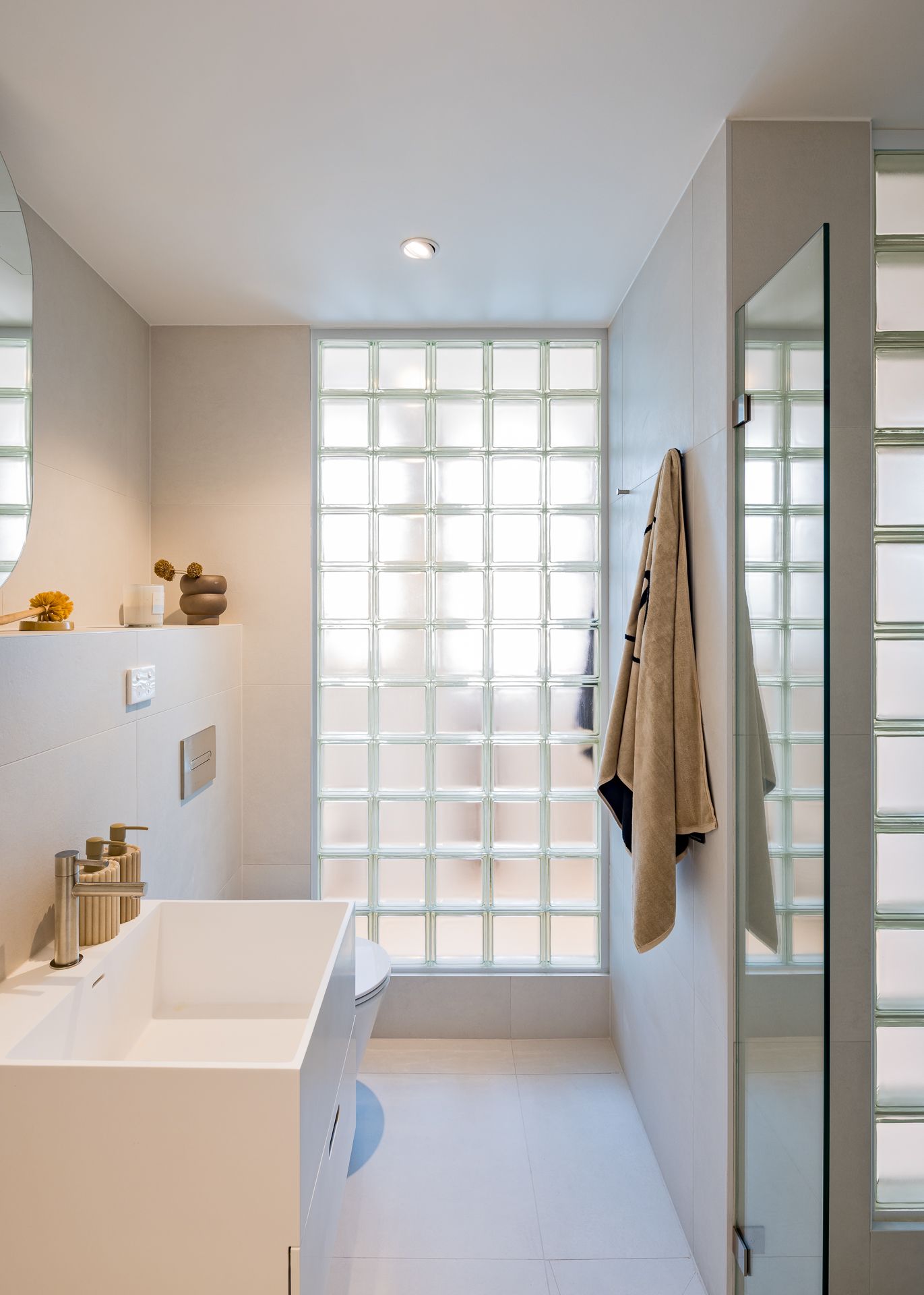 Modern bathroom with a white vanity basin, glass block wall, hanging towel and mirror in soft natural light.