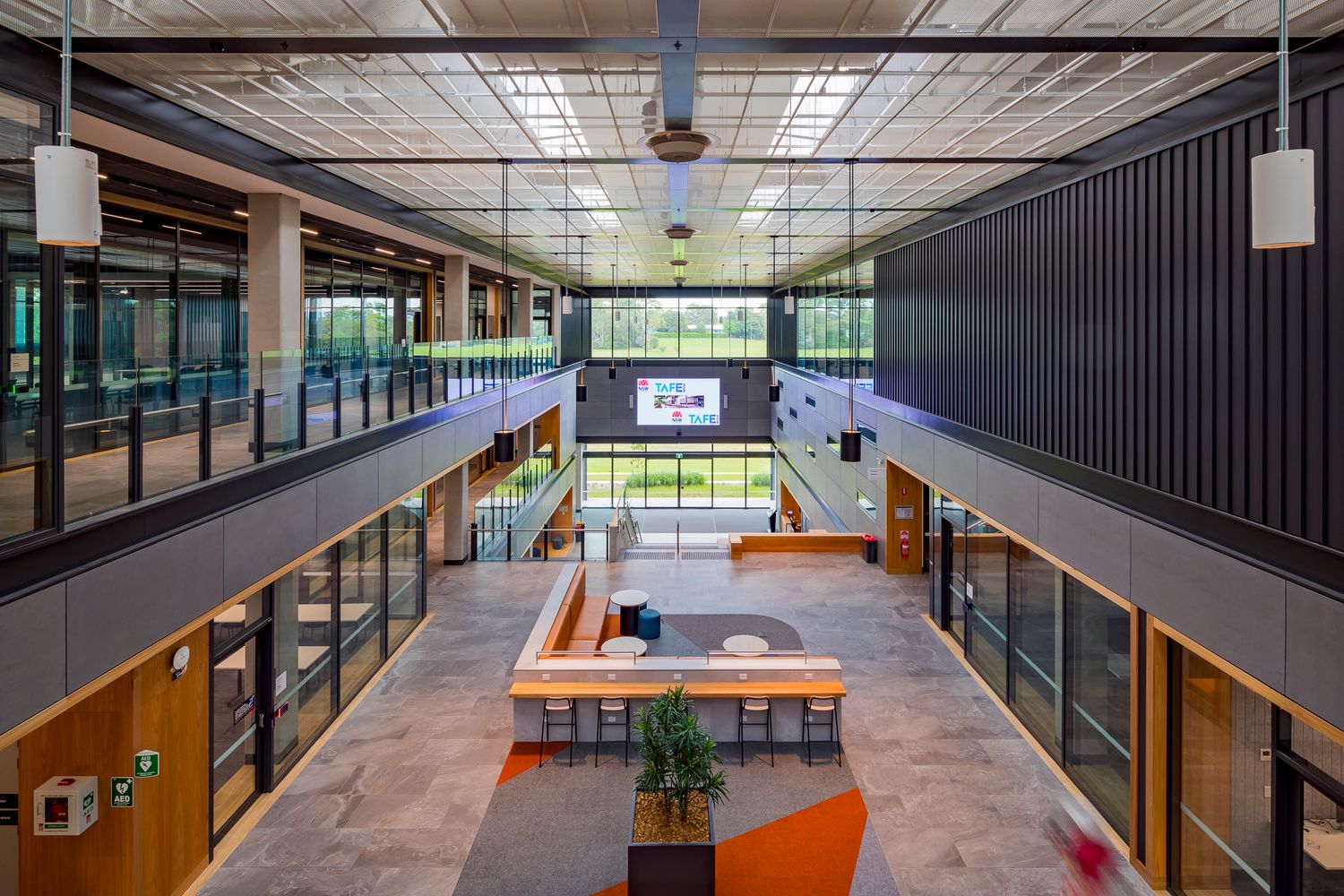 Open-plan atrium with timber bench seating, orange rug, and plants, framed by glass balconies and a skylit ceiling in a modern campus.