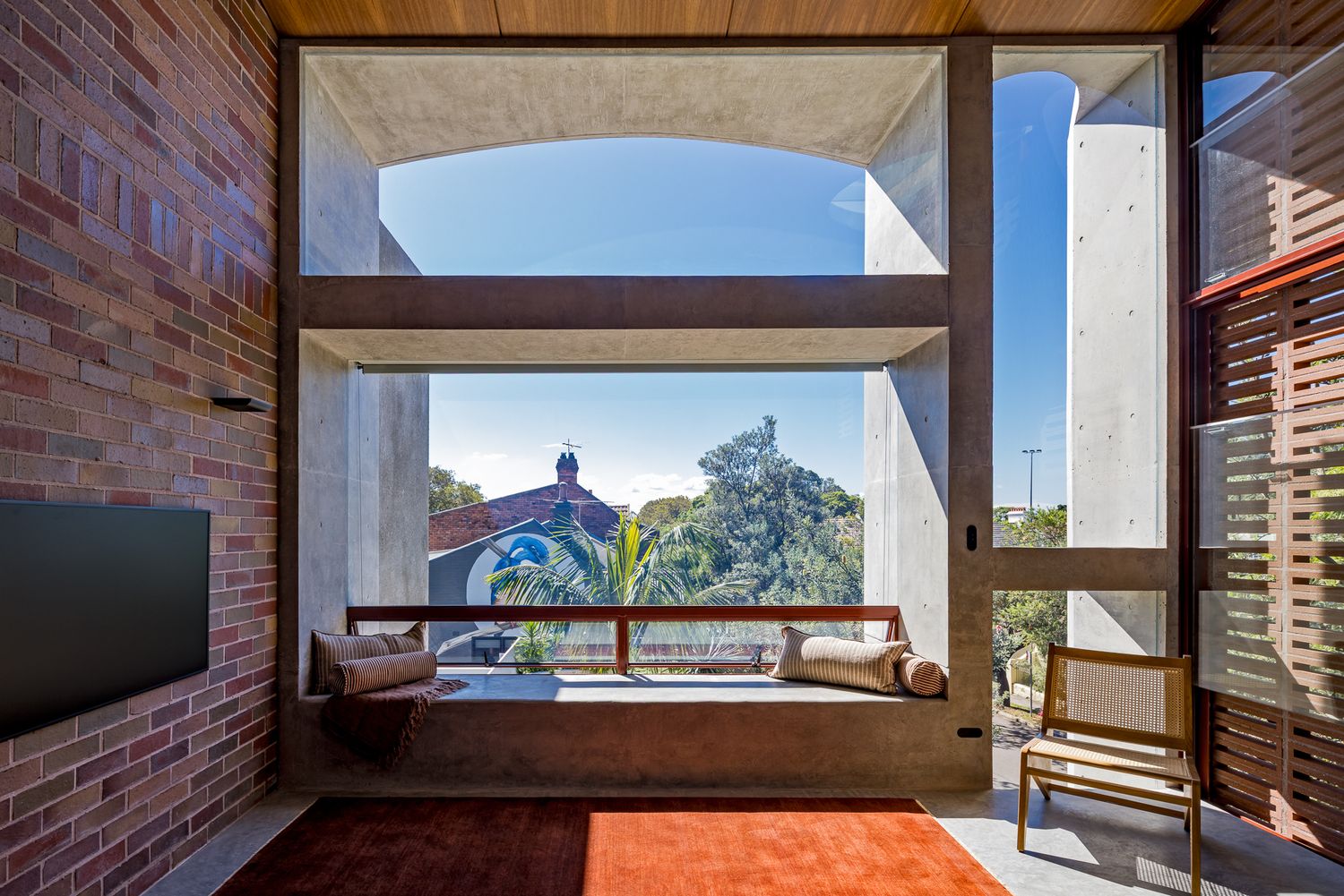 Arched concrete window framing a cushioned window seat and garden views, with a red rug, timber chair and brick wall.