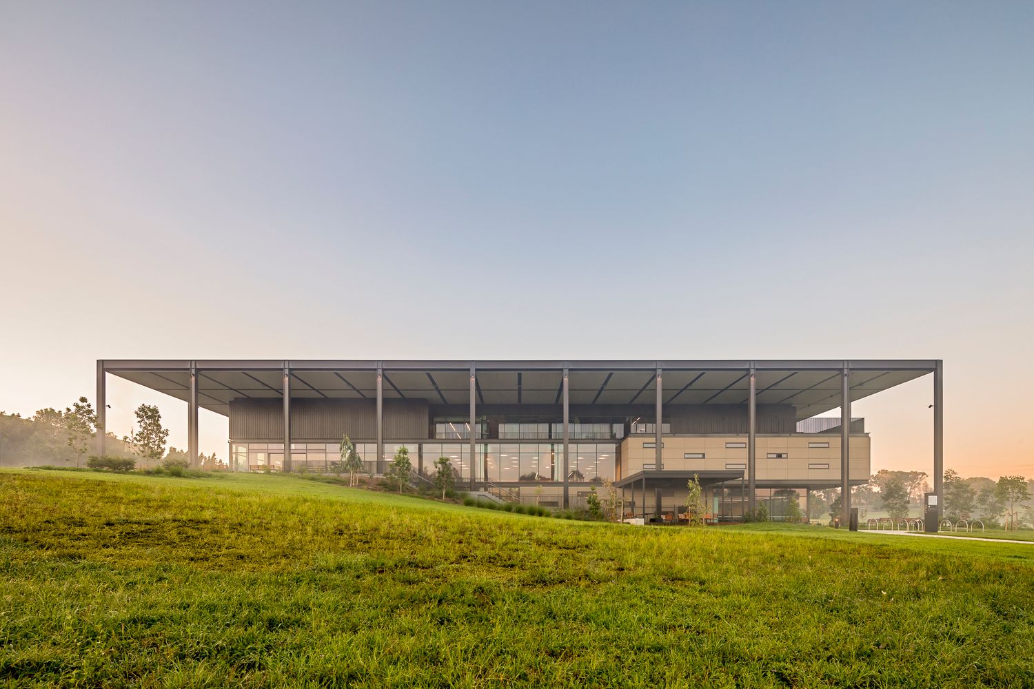Low-slung steel-framed building with a flat roof canopy rising above a grassy hill in soft morning mist.