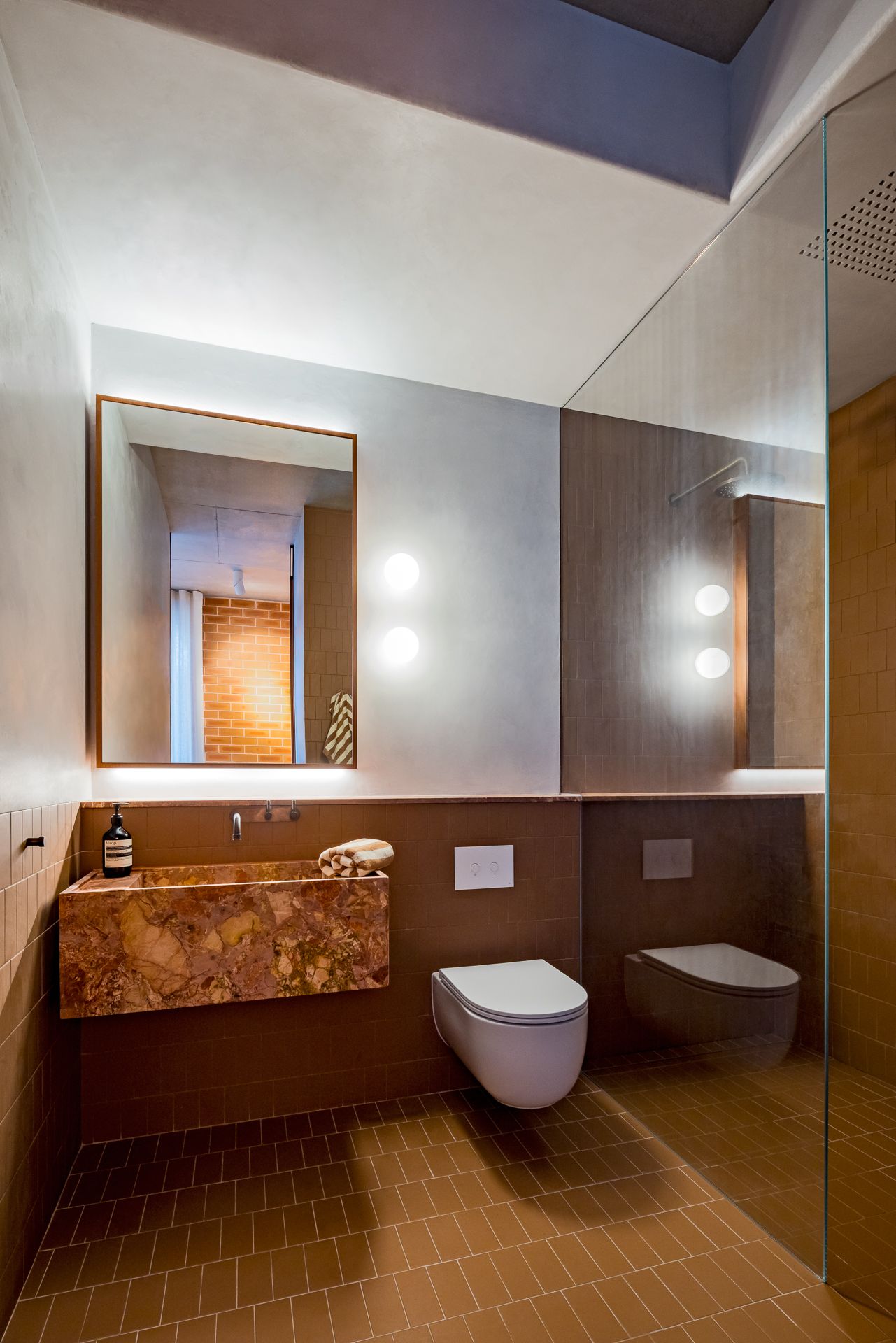 Modern bathroom with a marble vanity basin, backlit mirror, wall-hung toilet, terracotta floor tiles and a glass shower screen.