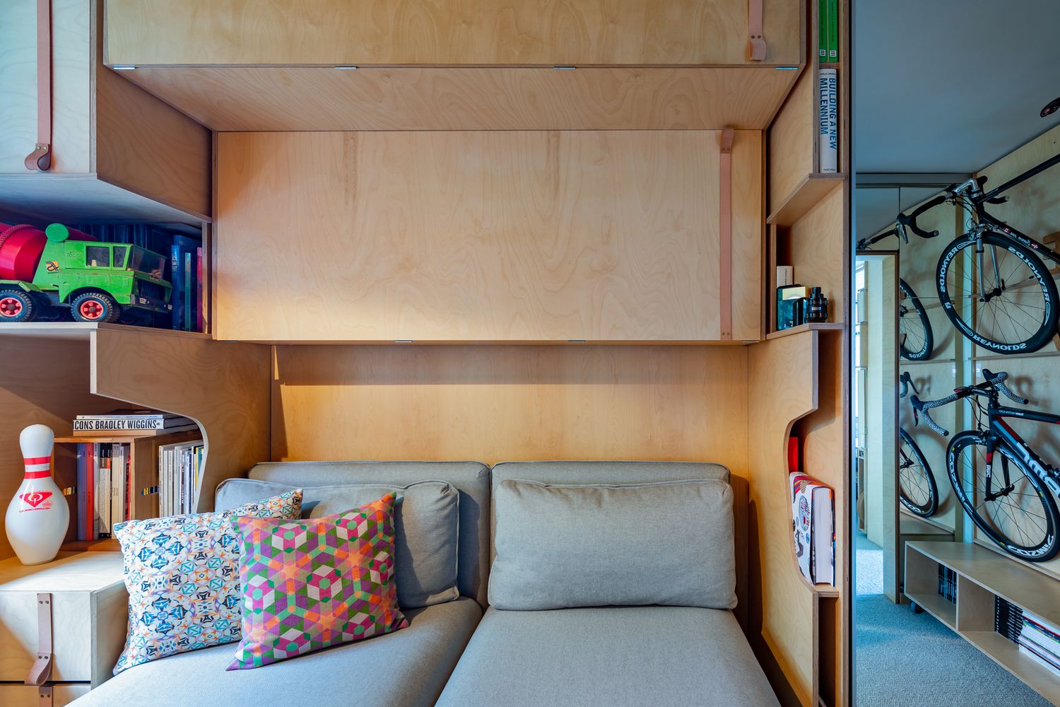Cozy plywood nook with a gray sofa, colorful cushions, toy truck and bowling pin on shelves, and bicycles visible through a mirrored door.