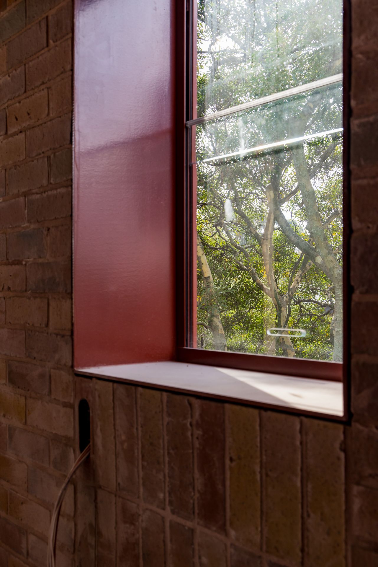 Deep red-framed window set into a brick wall with a sloped sill, looking out onto green trees and a bright sky.